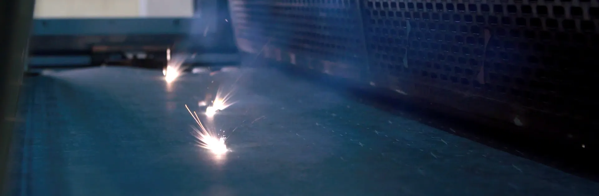 A close-up shot of an industrial laser cutting, showing multiple bright light points sparking as they interact with a dark blue metallic surface. The background features a perforated metal grate in a dimly lit, high-tech manufacturing environment.